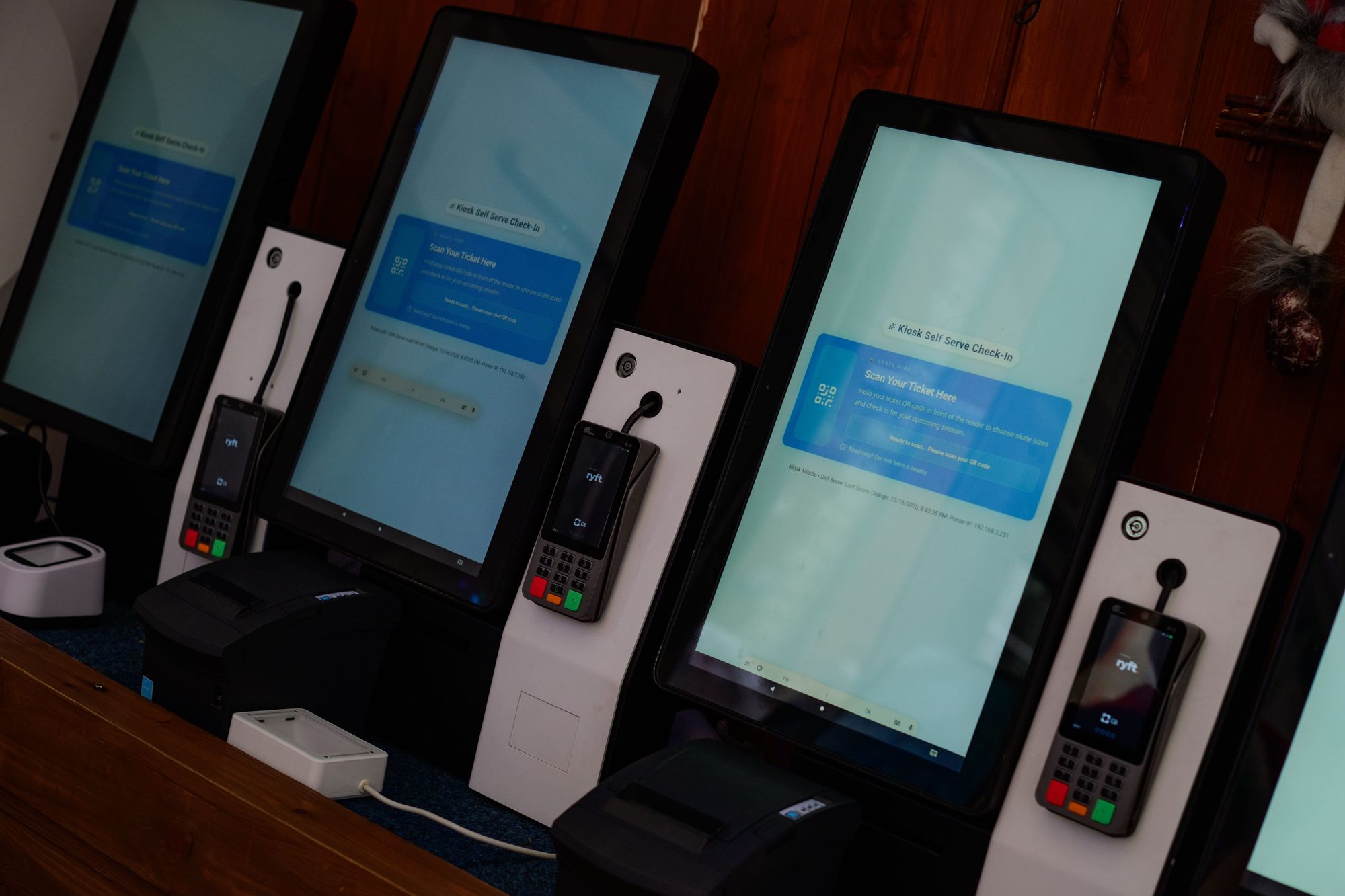Three self-service kiosks with portrait touchscreens, card terminals and receipt printers stand on a wooden counter.