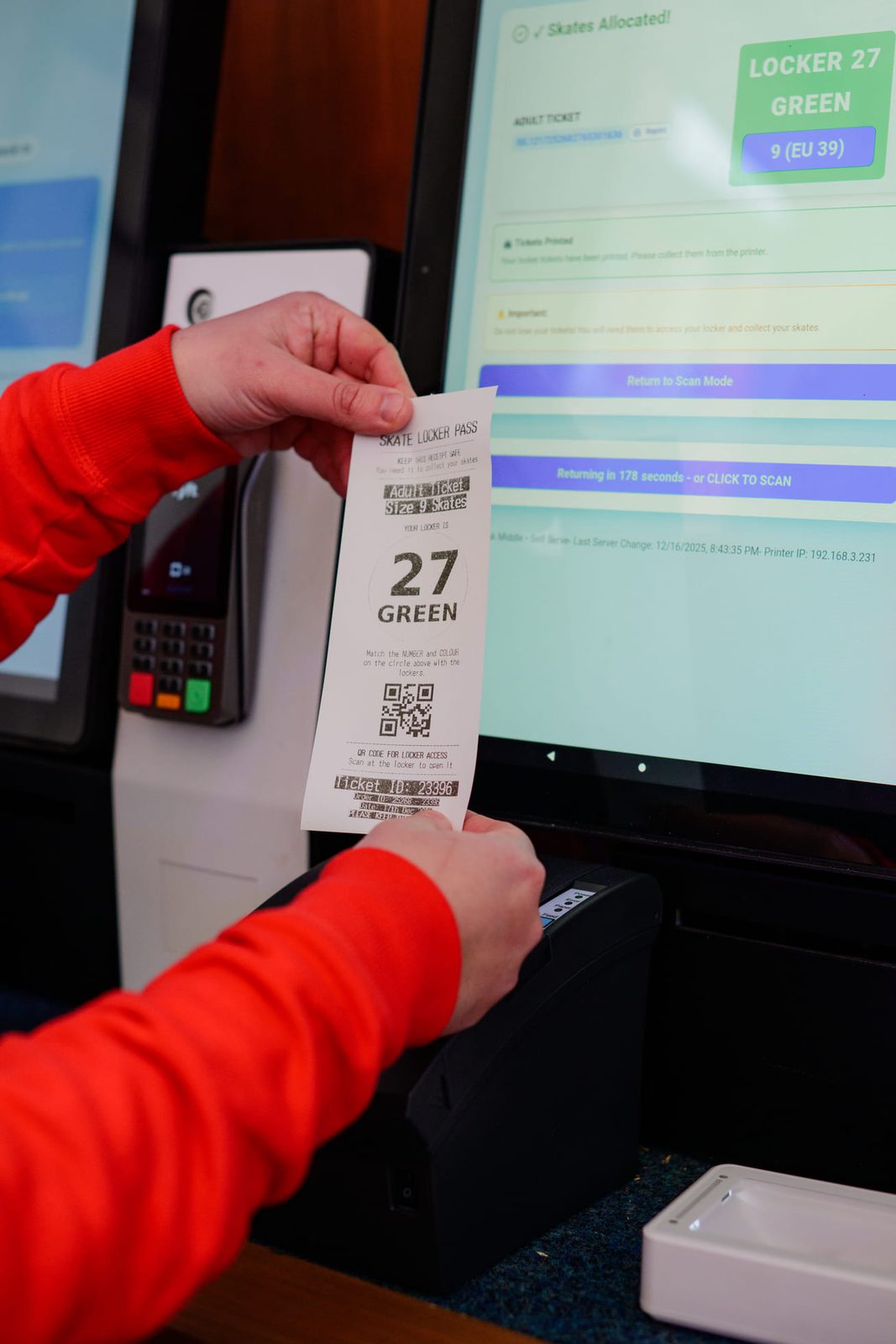 Hands take a printed locker pass from a kiosk printer below a screen that reads skates allocated, with a card terminal alongside.