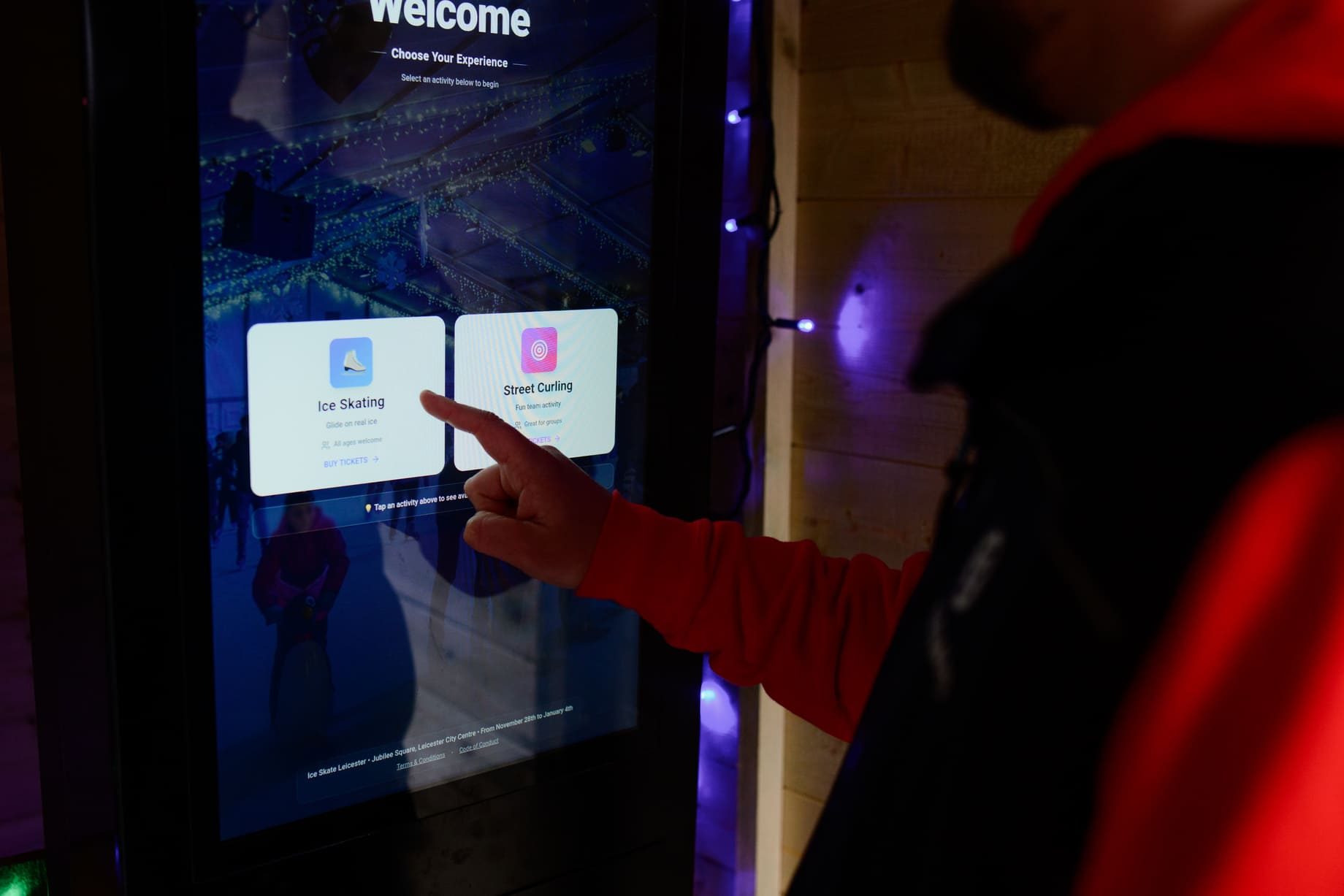 A finger taps a kiosk welcome screen to choose between two activities, with fairy lights on the wooden wall behind.