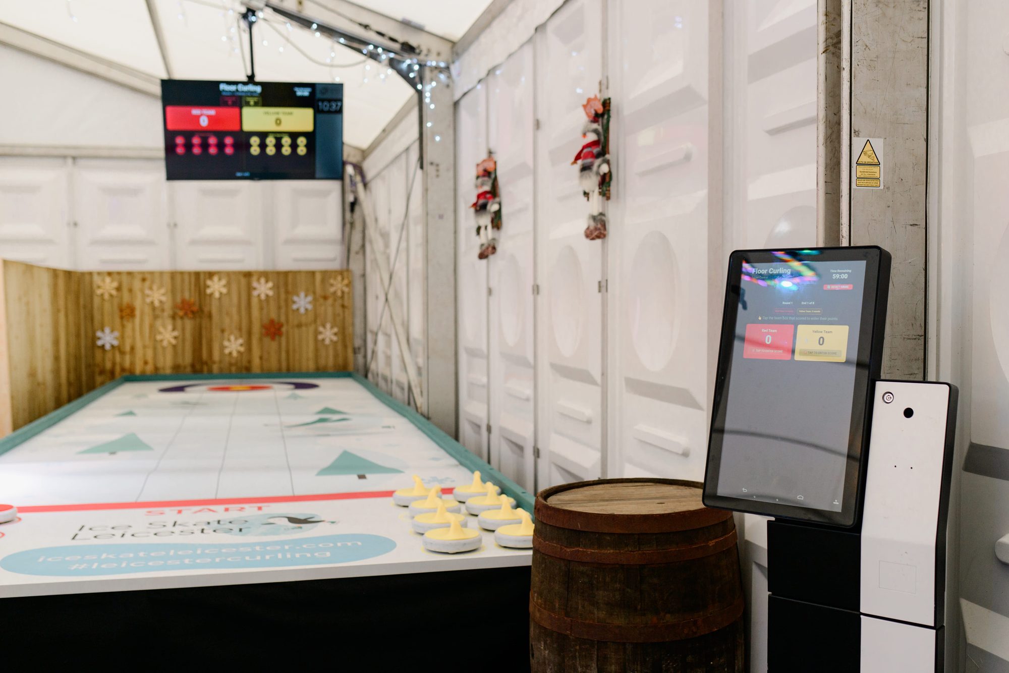A kiosk on a stand beside a curling lane with stones and a scoreboard screen, inside a marquee with container walls.